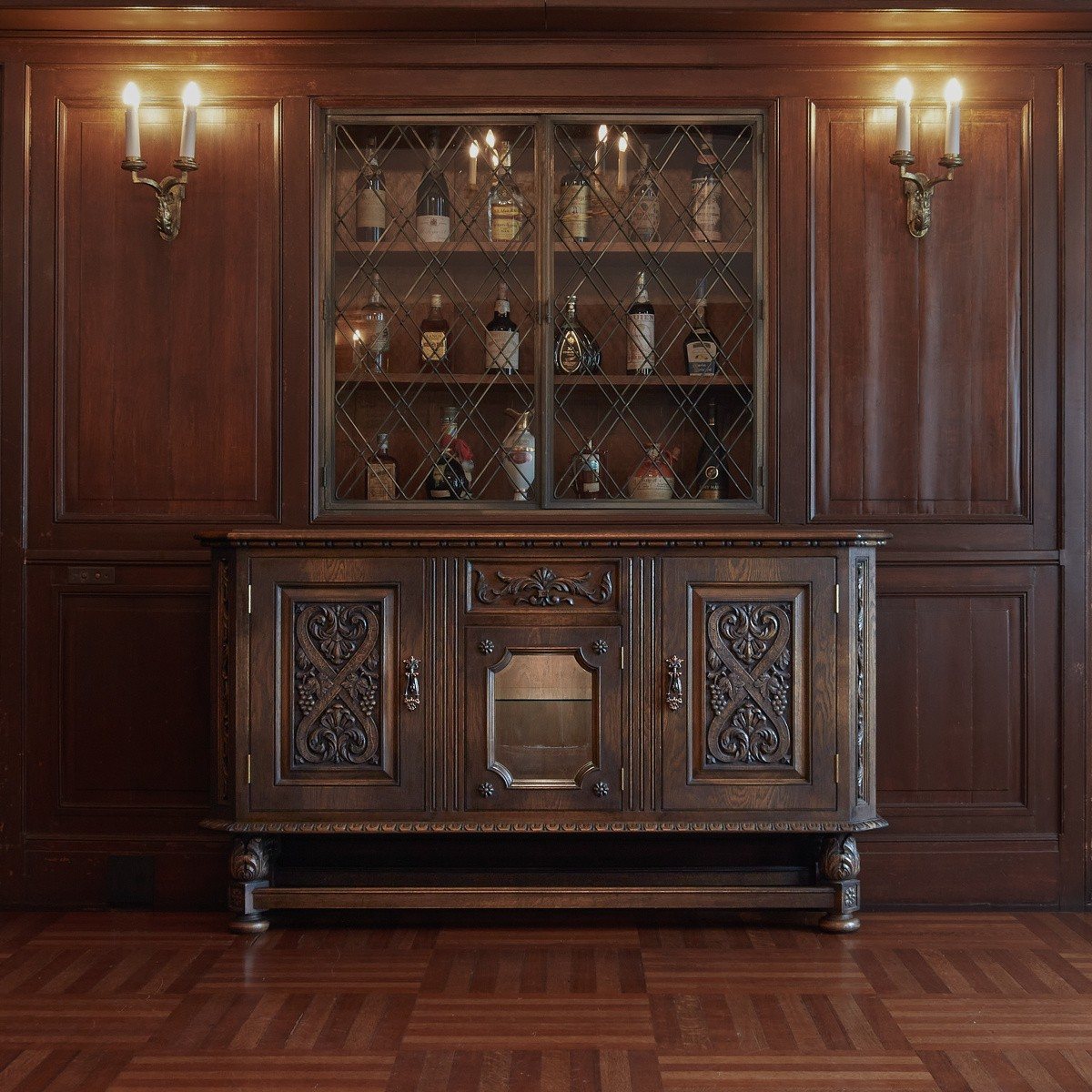 Carved wooden sideboard with a glazed upper cabinet, set against wood-panelled walls with wall lights